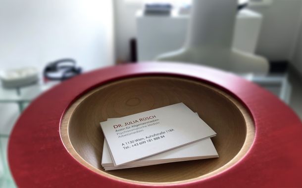 Stapel von Visitenkarten von Dr. Julia Rüsch, Fachärztin für Allgemeinmedizin und Psychosomatische Medizin, präsentiert in einer roten Schale auf einem modernen Glastisch in ihrer Wahlarztordination in Wien.